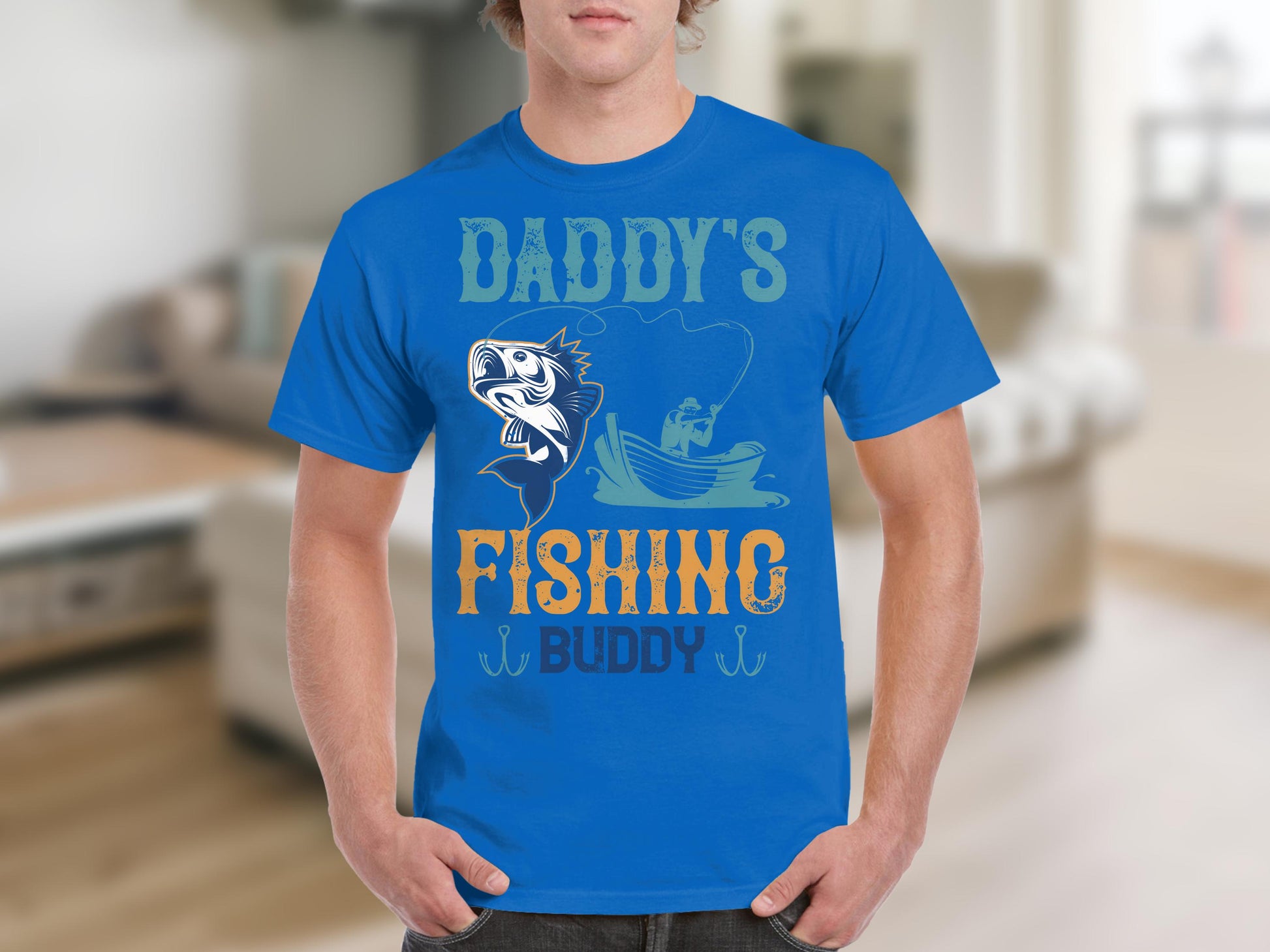 A person indoors wears a Garment Graphics blue t-shirt for fishing enthusiasts, featuring Daddys Fishing Buddy with a cartoon fish and someone in a boat. The softly blurred living room enhances the cozy vibe of this graphic print shirt.