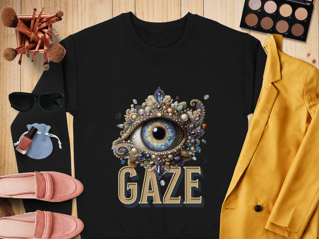A Garment Graphics black graphic sweatshirt featuring an ornate eye design above GAZE is displayed on a wooden surface. Its surrounded by makeup brushes, an eyeshadow palette, pink loafers, nail polish, sunglasses, and a yellow blazer, showcasing its versatile unisex sizing appeal.