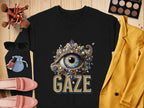 A Garment Graphics black graphic sweatshirt featuring an ornate eye design above GAZE is displayed on a wooden surface. Its surrounded by makeup brushes, an eyeshadow palette, pink loafers, nail polish, sunglasses, and a yellow blazer, showcasing its versatile unisex sizing appeal.