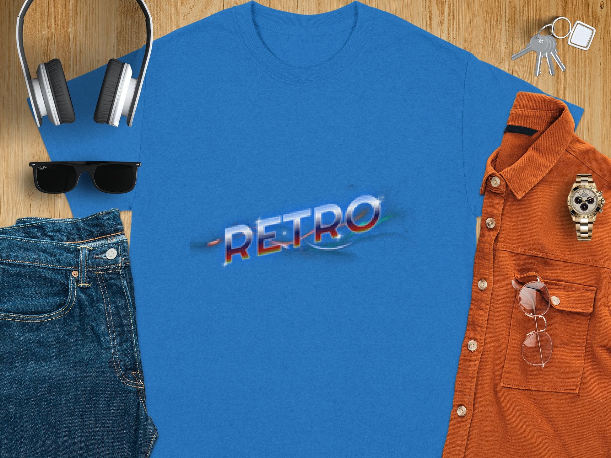 A Garment Graphics blue T-shirt featuring a retro design with Retro sits on a wooden surface, accompanied by headphones, sunglasses, denim jeans, an orange shirt holding glasses, a watch, and keys.