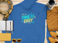 A blue Garment Graphics hoodie with Take It Easy in vibrant letters is styled on a wooden surface. It stands out among a plaid scarf, yellow clutch, sunglasses, coffee cup, quilted jacket, and yellow shoes—ideal for a cozy fashion statement with its bright blue and yellow look.