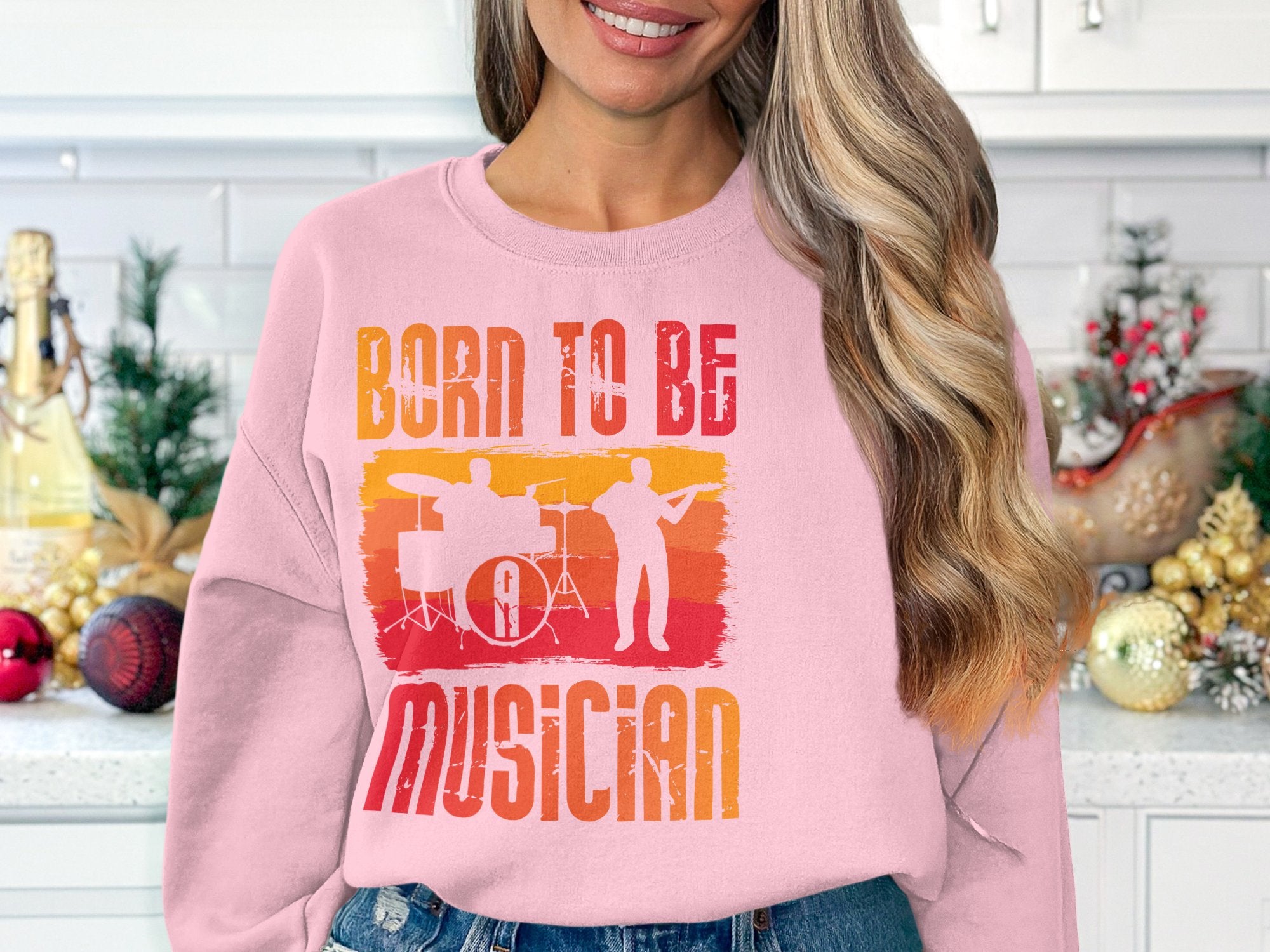 A woman with long hair dons a Garment Graphics pink sweatshirt featuring Born to Be Musician and musician silhouettes. She stands in a festively decorated kitchen, ideal for music enthusiasts. Available in unisex sizes, it suits anyone embracing their musical spirit.