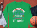 A Garment Graphics green fishing sweatshirt sits on a brown surface, displaying the text Id rather have a bad day fishing than a good day at work. Perfect for enthusiasts, its next to a water bottle, towel, black shorts, and basketball. Unisex sizing ensures ultimate comfort.