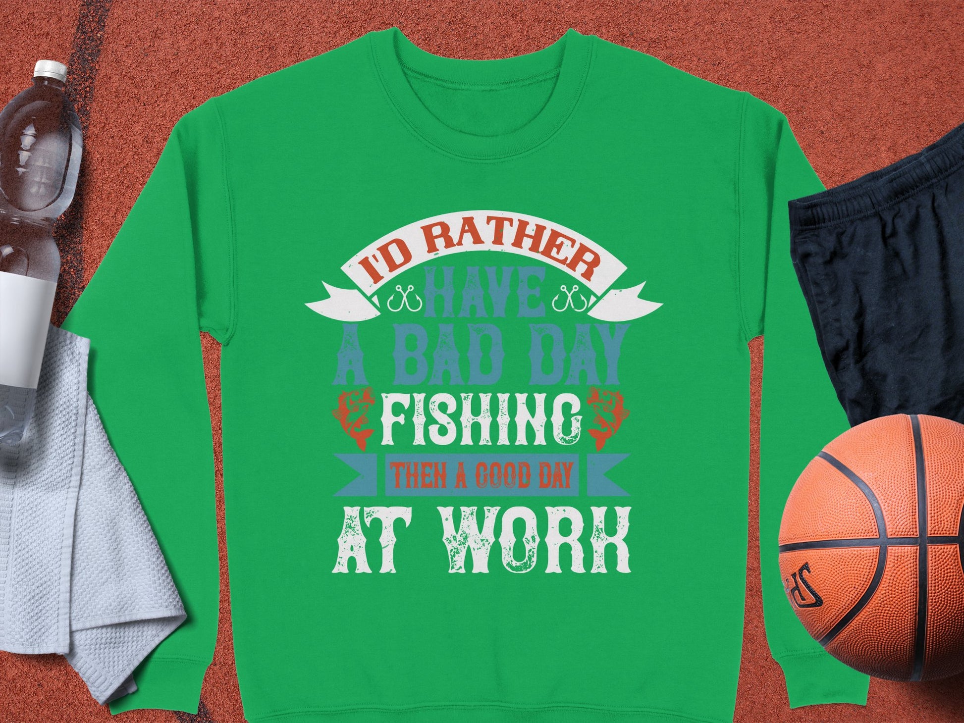 A Garment Graphics green fishing sweatshirt sits on a brown surface, displaying the text Id rather have a bad day fishing than a good day at work. Perfect for enthusiasts, its next to a water bottle, towel, black shorts, and basketball. Unisex sizing ensures ultimate comfort.