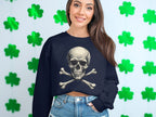 A person in Garment Graphics unisex black sweater featuring a skull and crossbones design stands against green shamrocks. Their long brown hair flows freely as they smile, effortlessly chic with denim jeans.