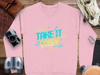 A unisex pink sweatshirt by Garment Graphics showcases the Take It Easy graphic in vibrant blue and yellow with musical notes and arrows. Its comfortably styled on a wooden surface alongside a camera, binoculars, gloves, a bag, and a carabiner.