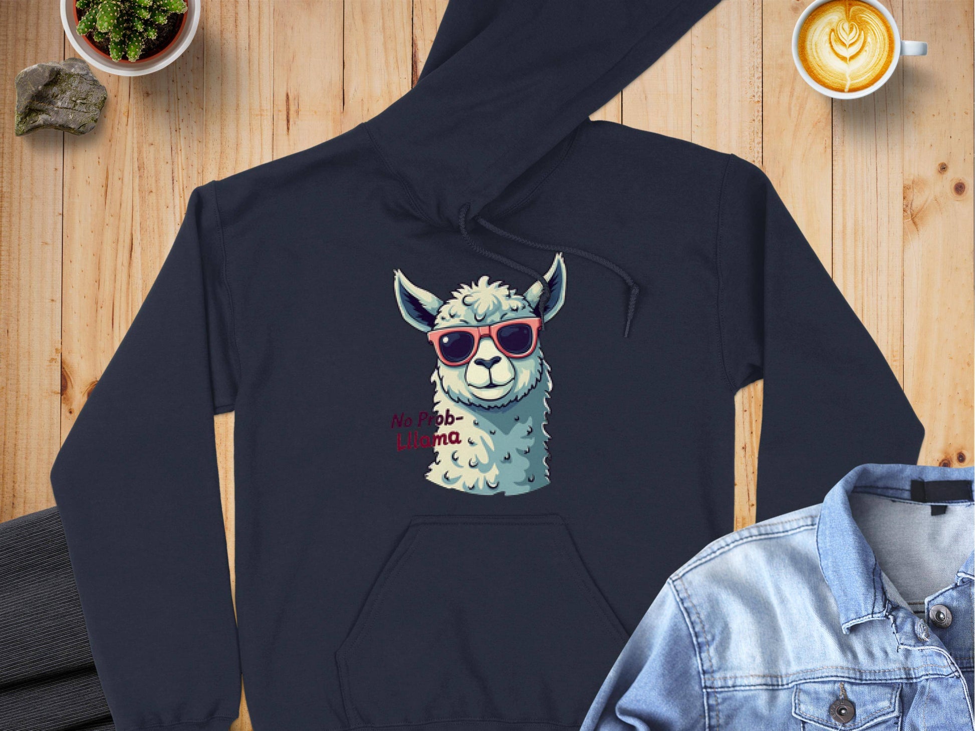 A black llama hoodie by Garment Graphics with sunglasses and No Prob-Llama text is on a wooden surface, paired with a denim jacket, latte with art, and two potted plants—a perfect quirky style addition to your classic fit wardrobe.