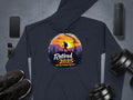 Navy blue hiking hoodie by Garment Graphics featuring a hiker silhouette with Retired 2025 Not My Problem Anymore on a sunset mountain design, surrounded by items like a phone, smartwatch, Bluetooth speaker, dumbbells, and black sneakers.