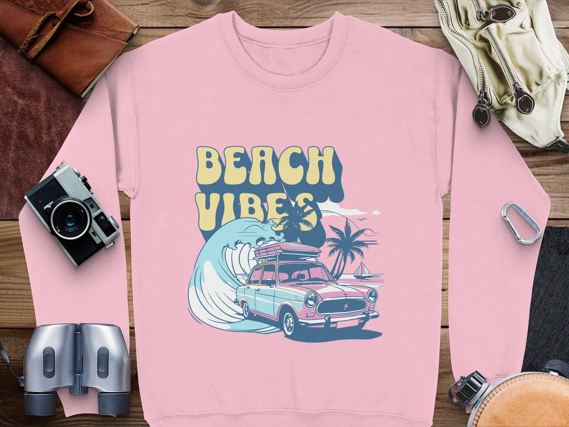 Garment Graphics’ pink sweatshirt boasts a retro design of a classic car with surfboards, palm trees, and a wave. The BEACH VIBES text highlights the graphic, adding to its charm. Surrounded by camera gear, leather items, and a carabiner on wood, it exudes undeniable nostalgia.