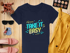 A flat lay features a Garment Graphics navy T-shirt with vibrant TAKE IT EASY text. Surrounding items include a mustard yellow blazer, pink loafers, sunglasses, makeup brushes, an eyeshadow palette, nail polish, and earrings on a wooden surface.