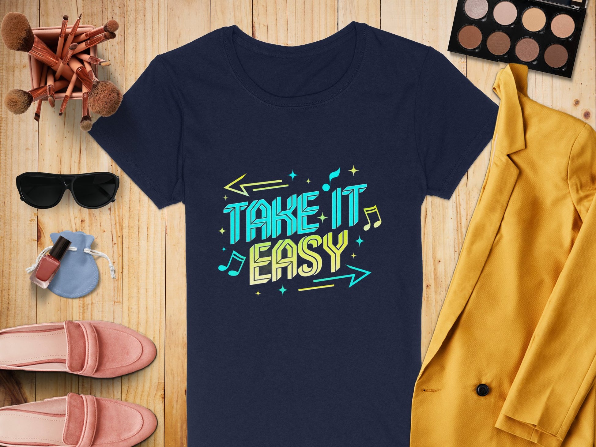 A flat lay features a Garment Graphics navy T-shirt with vibrant TAKE IT EASY text. Surrounding items include a mustard yellow blazer, pink loafers, sunglasses, makeup brushes, an eyeshadow palette, nail polish, and earrings on a wooden surface.