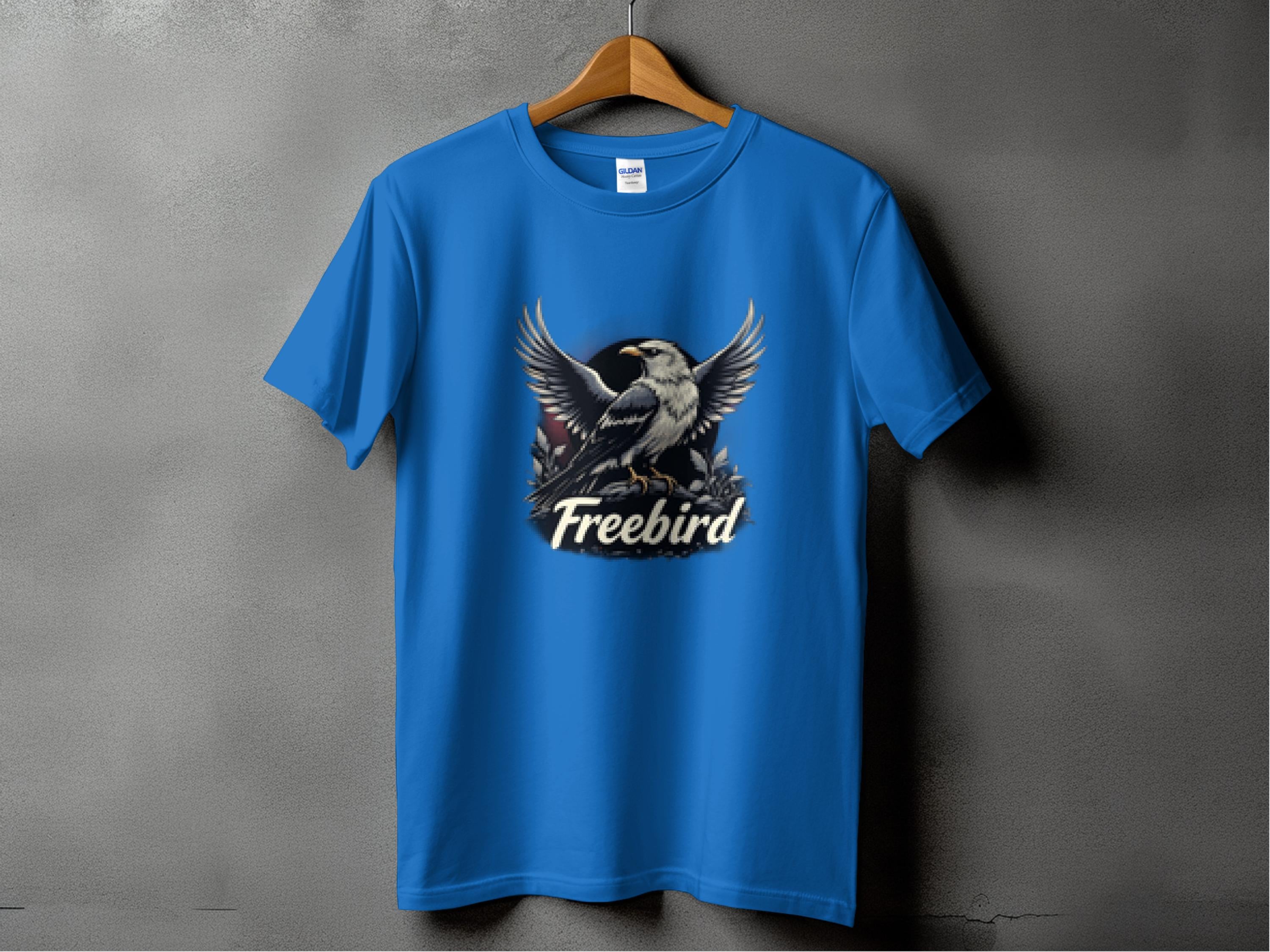 A blue, 100% cotton T-shirt by Garment Graphics hangs on a wooden hanger against a gray backdrop. It showcases a bold graphic of a bird with outstretched wings and Freebird beneath, making it the perfect eagle T-shirt for any casual day.