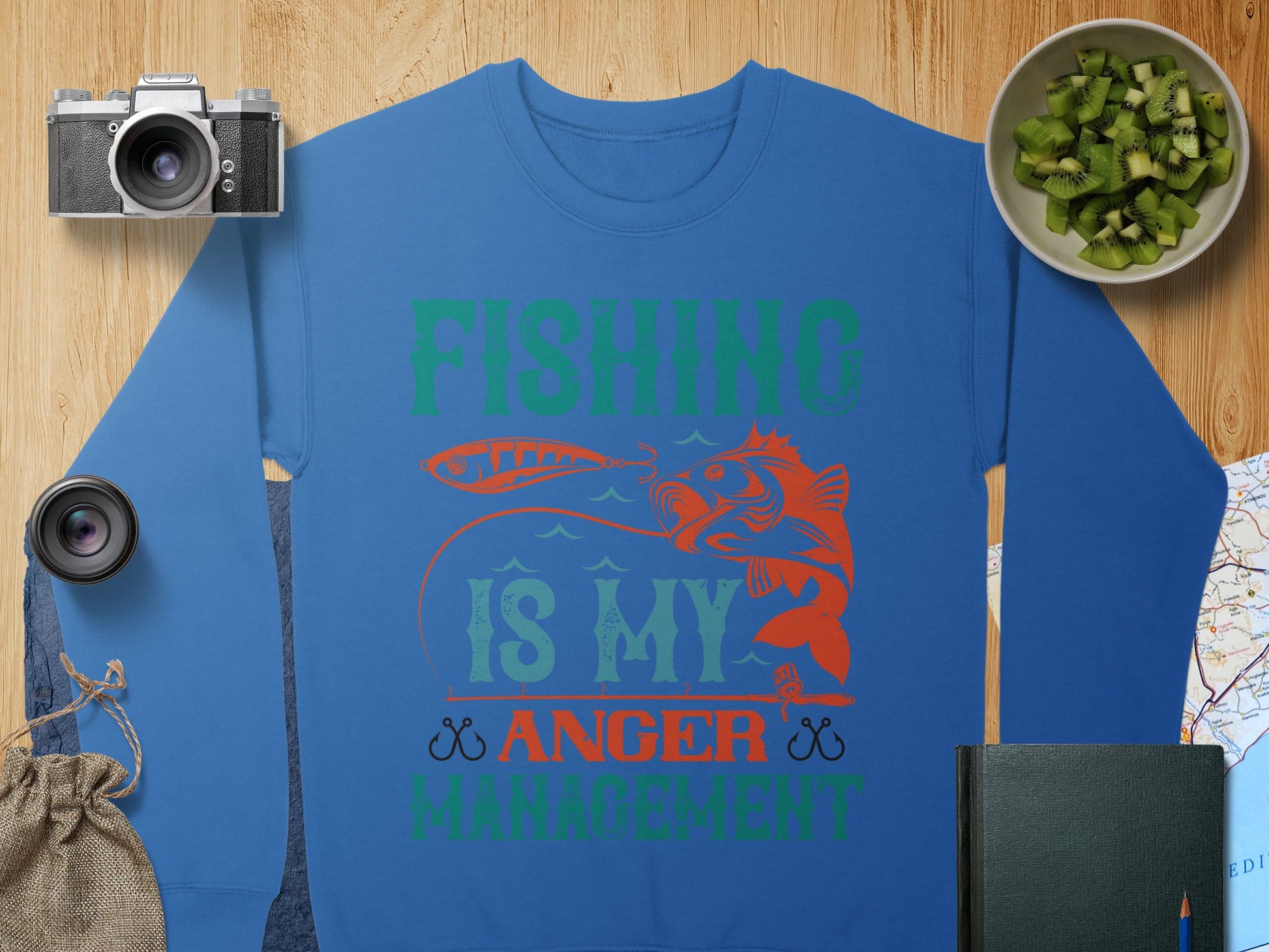 The Garment Graphics blue sweatshirt showcases a unique design with Fishing Is My Anger Management in green and orange, featuring a fish holding a lure. It includes kiwi slices, a camera lens, and a map for an adventurous vibe, ideal for fishing enthusiasts.