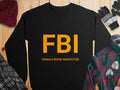 A black Garment Graphics sweatshirt with FBI in bold yellow and Female Boob Inspector beneath, displayed on a wooden surface alongside a hat, gloves, plaid shirt, and jacket. Unisex-sized with a graphic print design.