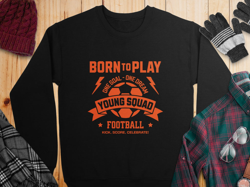 A unisex black football sweatshirt from Garment Graphics with orange graphics and text: Born to Play, a soccer ball, Young Squad Football, plus the slogans One Goal - One Dream and Kick. Score. Celebrate! showcased on wood with winter accessories nearby.