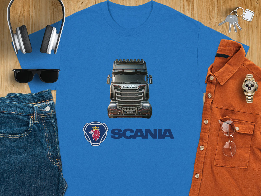 A person wears a Garment Graphics white T-shirt with a black Scania truck, ideal for truck lovers. The iconic Scania logo and name are beneath the image on a grassy field background.