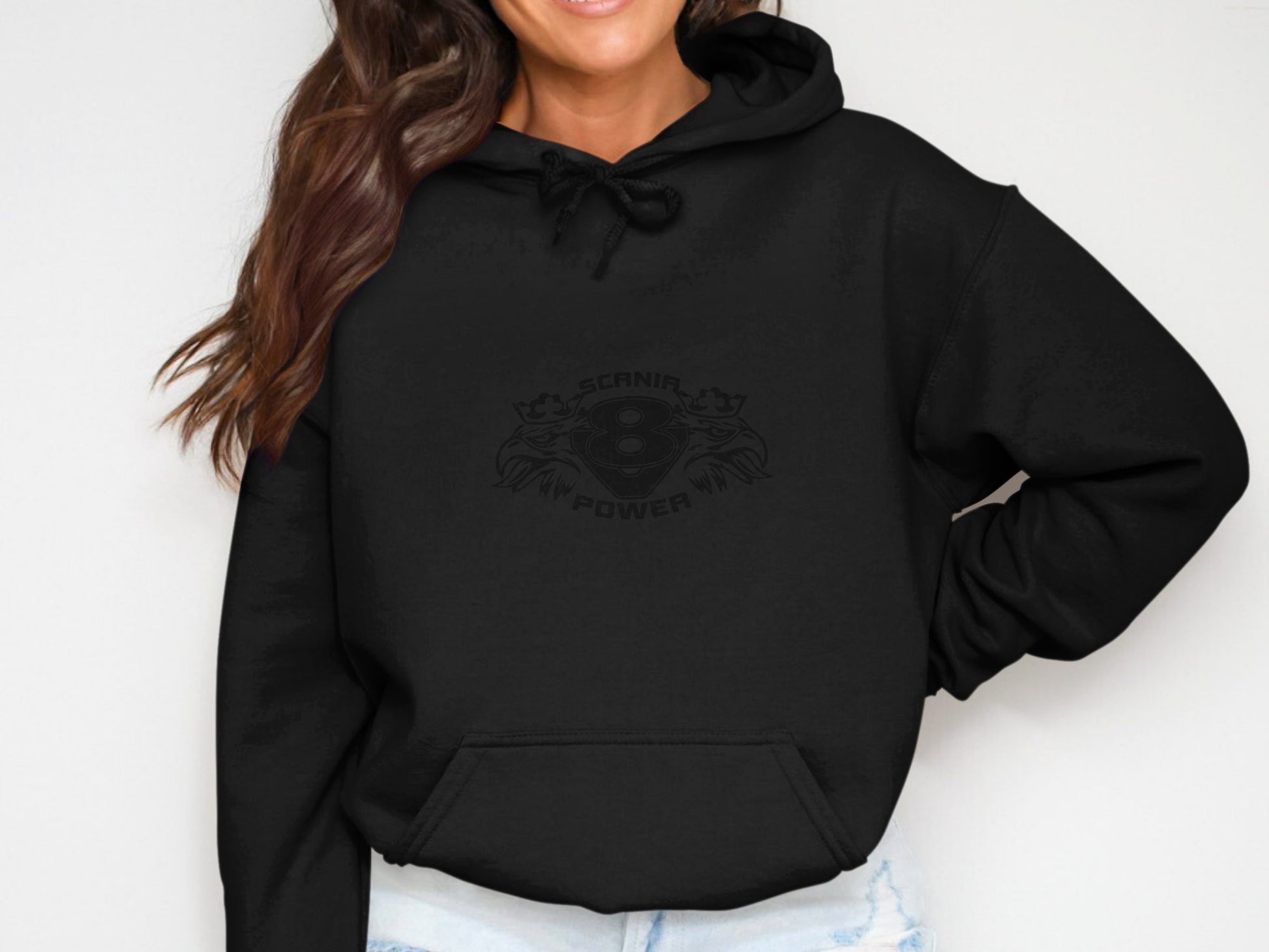 A person is wearing Garment Graphics Scania Power Eight Hoodie in black, showcasing a bold front design, drawstring neck, and large front pocket. With long wavy hair, they pose with one hand on their hip and are perfectly paired with classic fit light blue jeans.