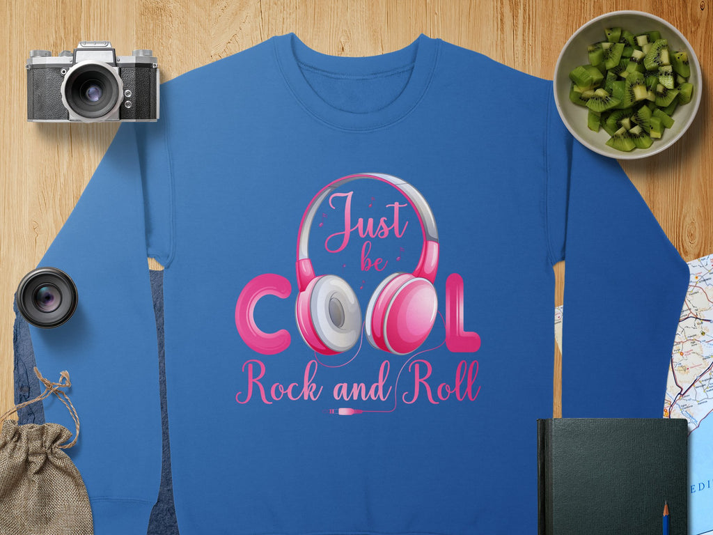 Garment Graphics blue sweatshirt showcases pink and gray rock & roll headphones with Just be Cool Rock and Roll text. Paired with a camera, lens, chopped kiwi bowl, journal, map, and a small bag—perfect for any fashion enthusiast.