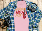 A unique pink Garment Graphics T-shirt featuring Music Never Sleeps and a guitar graphic is displayed on a wooden surface, complemented by a blue and green plaid shirt, headphones, a camera, and a white takeaway coffee cup—perfect for guitar T-shirt enthusiasts.