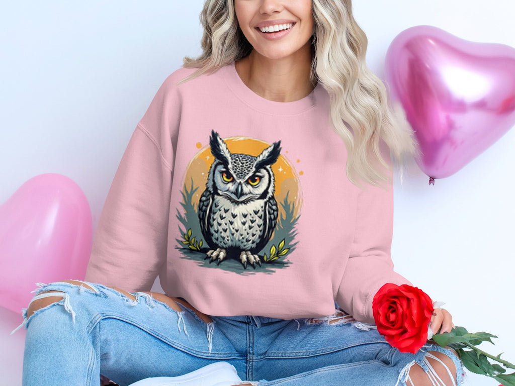 A person with long, wavy hair sits cross-legged wearing a unisex pink owl-design sweatshirt from Garment Graphics. They hold a red rose as two pink heart-shaped balloons float partially visible in the background.