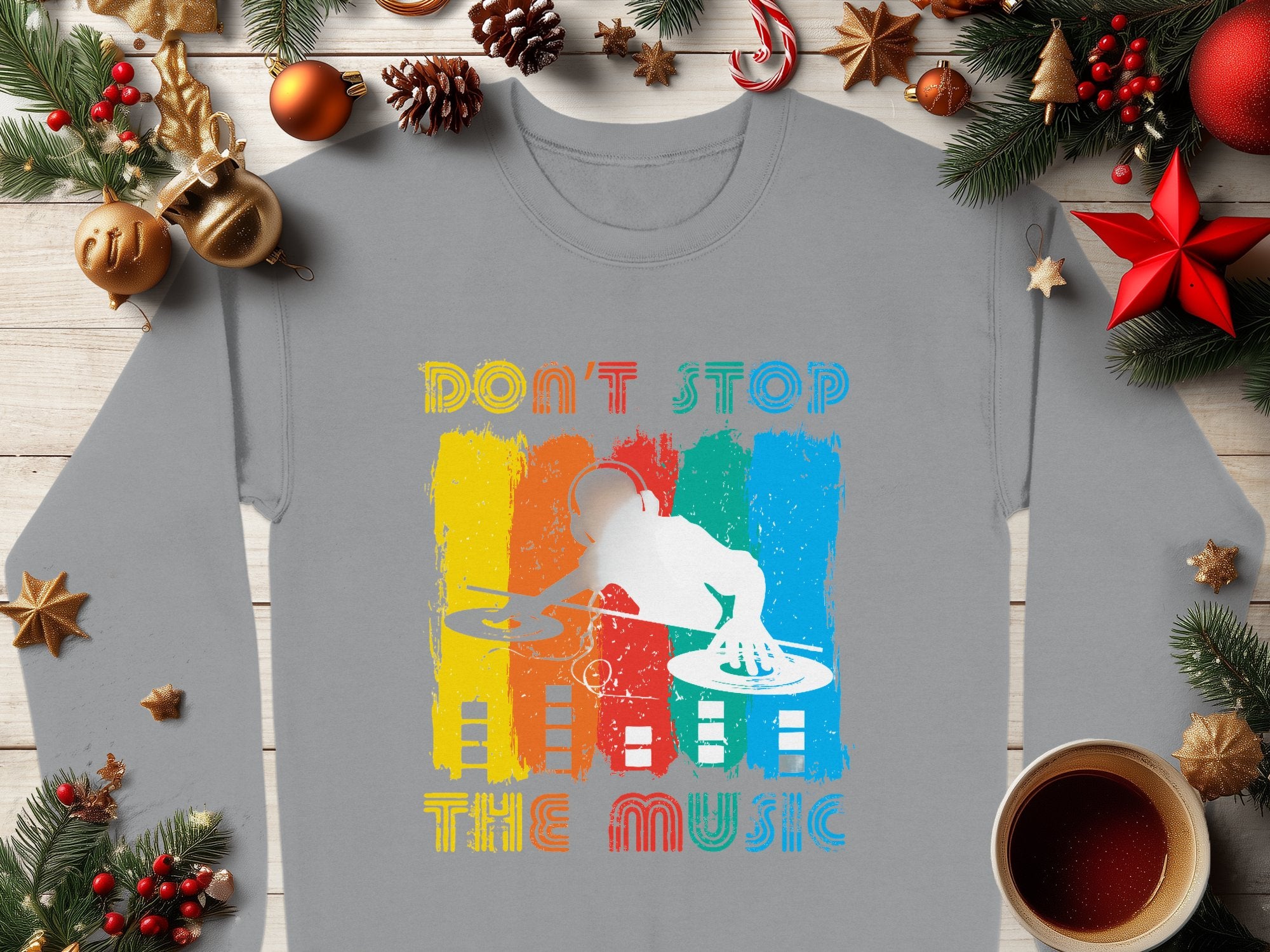 Garment Graphics offers a unisex gray shirt ideal for music enthusiasts, featuring a DJ silhouette with turntables set against vibrant orange, yellow, blue, and red vertical stripes. The shirt reads Dont Stop the Music and is styled with holiday decor and a cozy cup of coffee.