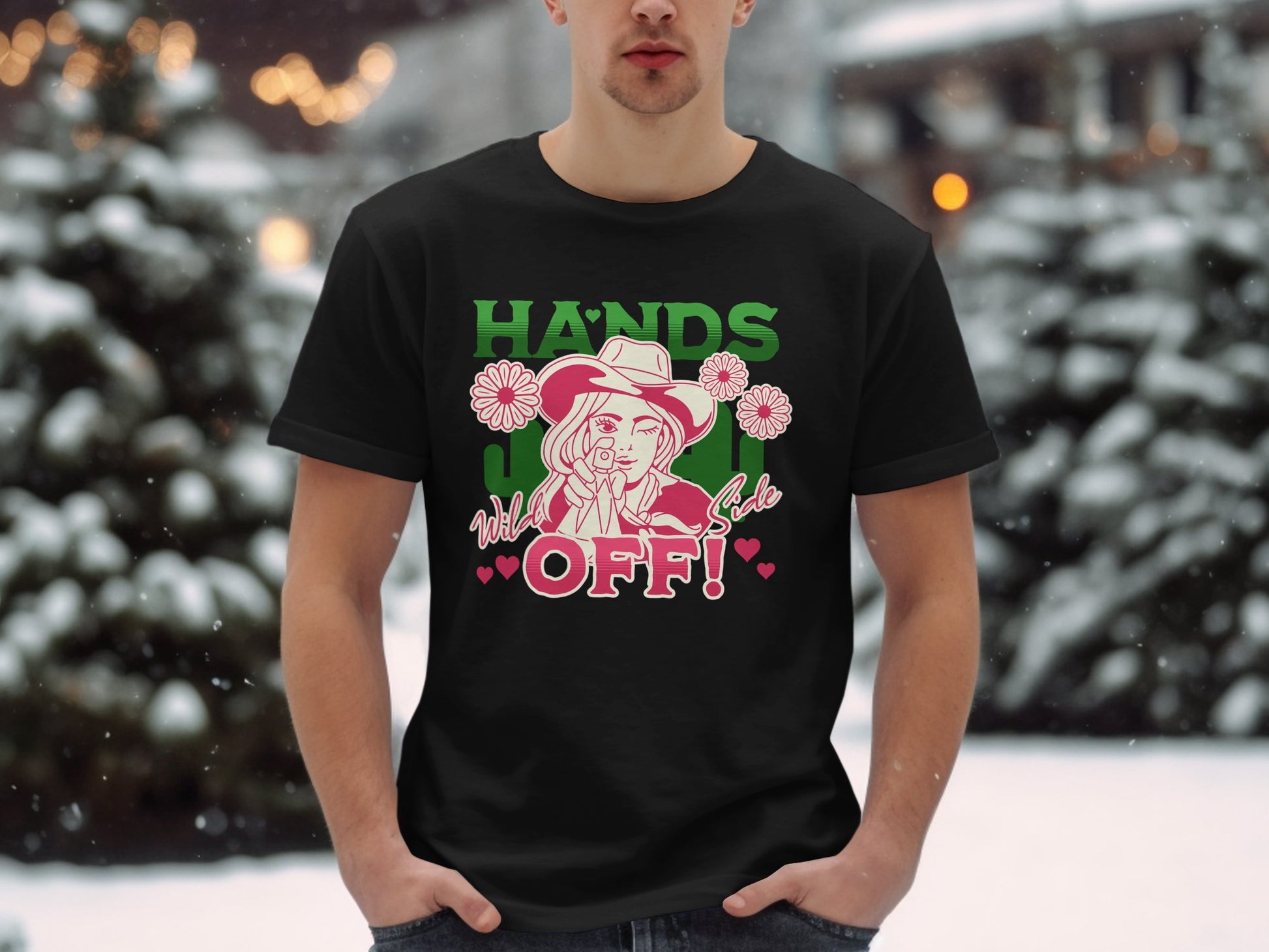 A person stands in the snow wearing a Garment Graphics classic fit black T-shirt. The shirt features a cartoon figure holding a rose in their mouth with HANDS OFF! printed on the front. Snow-covered trees create a serene backdrop for this eye-catching Hands Off T-Shirt.