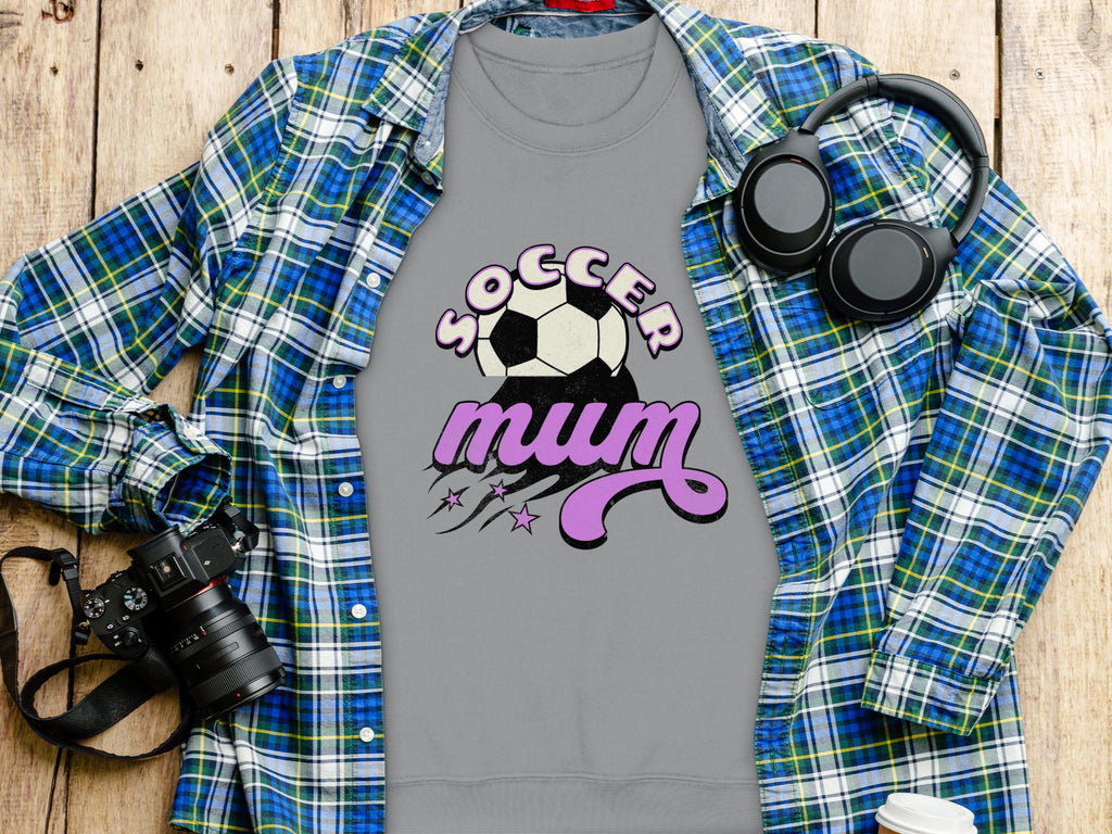A Garment Graphics gray shirt with Soccer Mum in bold and a vintage soccer ball graphic lies on a wooden surface. Nearby are a blue plaid shirt, headphones, camera, and takeout coffee cup, creating an adventurous everyday vibe.