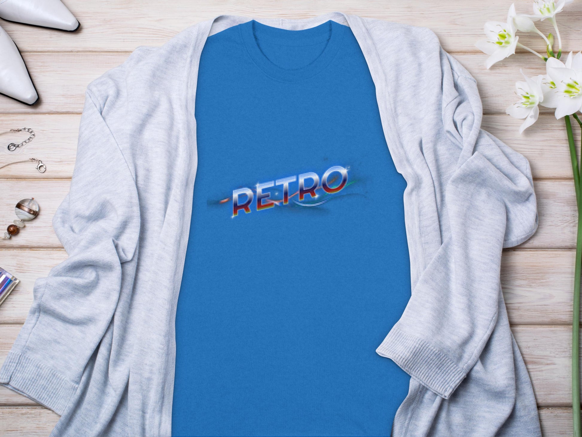 A Garment Graphics blue T-shirt with a Retro Graphic Design in colorful font rests on wood, exuding Vintage Charm. Partially covered by a light gray cardigan, it offers a classic fit. White flowers and small accessories enhance the scenes elegance.