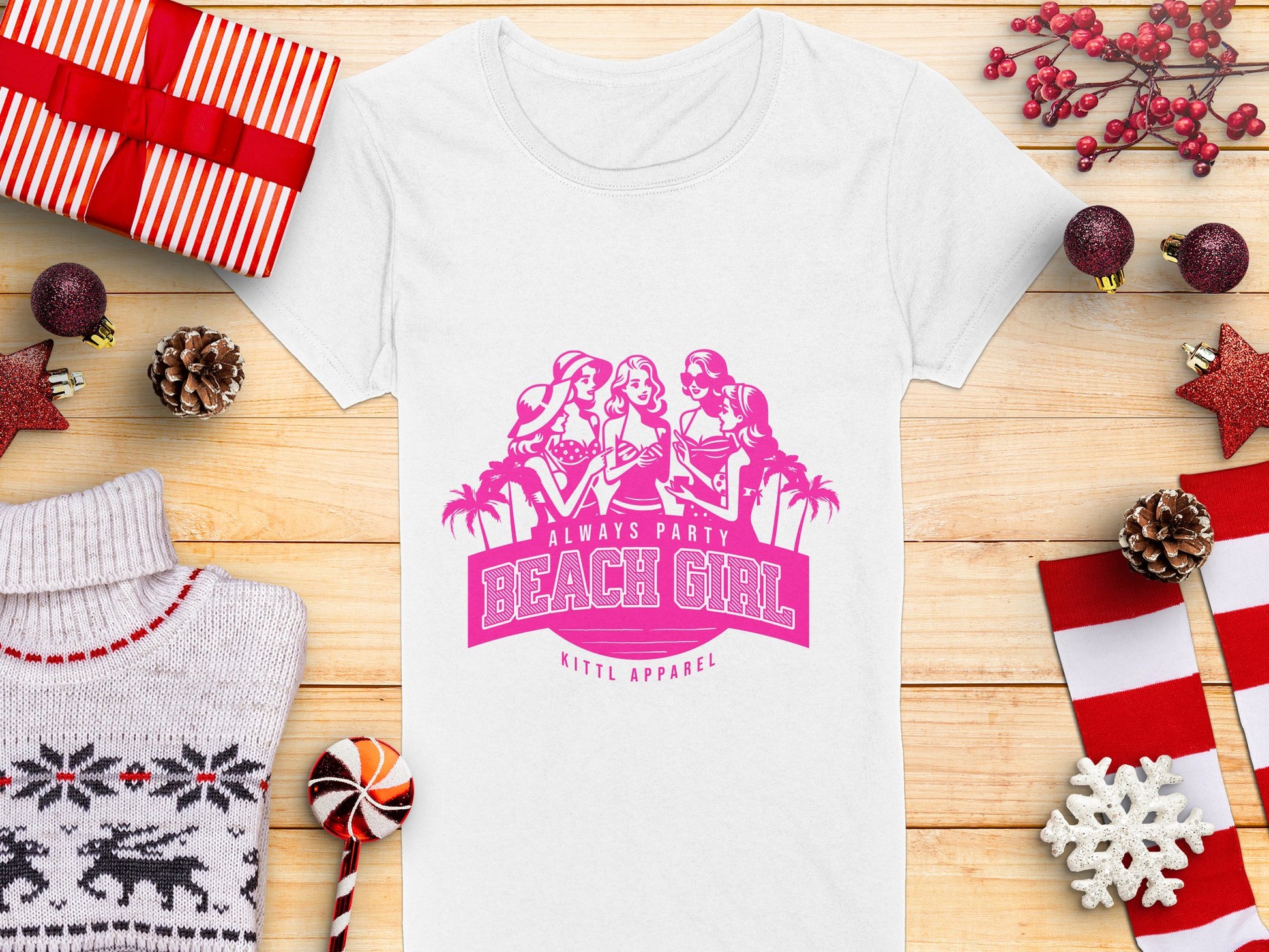 A white semi-fitted womens tee, branded Garment Graphics, displays a pink graphic of three women, palm trees, and the text Always Party Beach Girl above Kittl Apparel. The t-shirt is placed on a wooden surface with gifts, pine cones, berries, a candy cane, and a sweater.