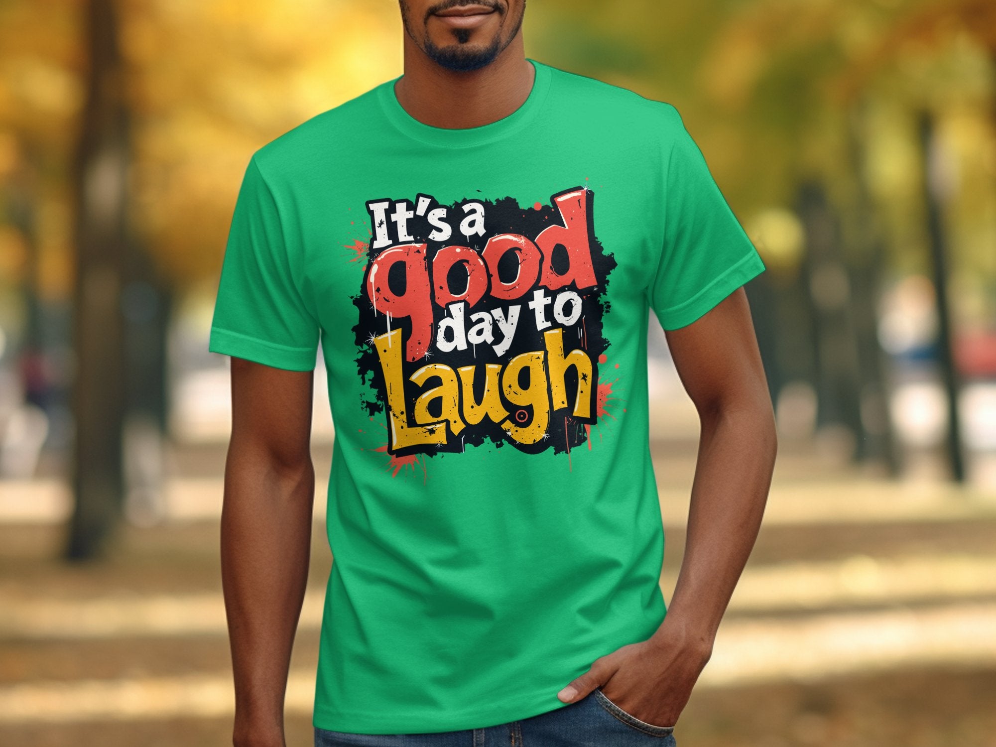 A person wears a bright green Garment Graphics Its a Good Day to Laugh T-shirt, showcasing bold, colorful letters. The backdrop displays blurred autumn trees and a leaf-covered path.