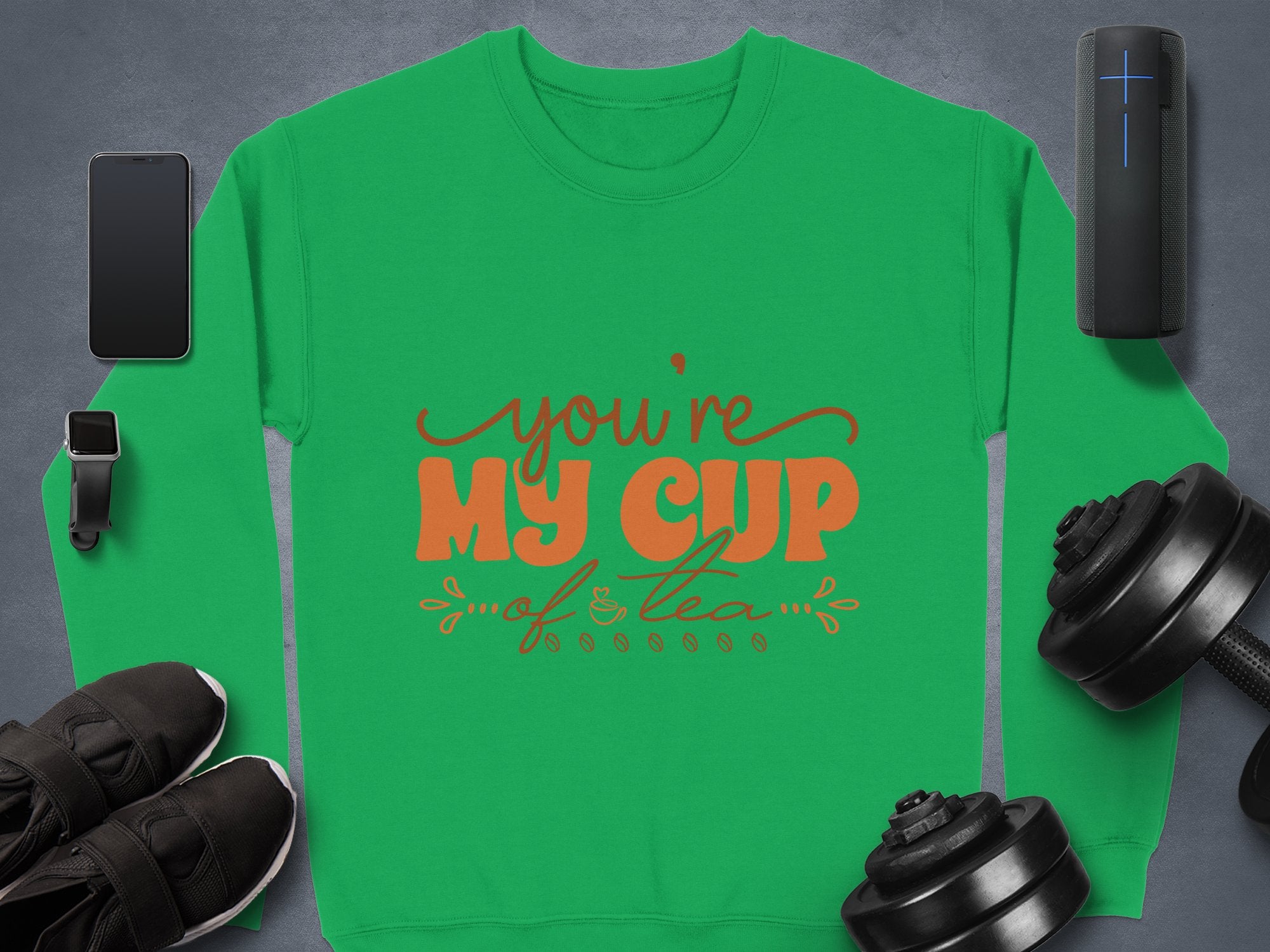 The Garment Graphics cozy unisex green sweatshirt features Youre My Cup of Tea text in vibrant orange and red, stylishly displayed with a smartphone, smartwatch, Bluetooth speaker, sneakers, and dumbbells on a sleek gray surface.