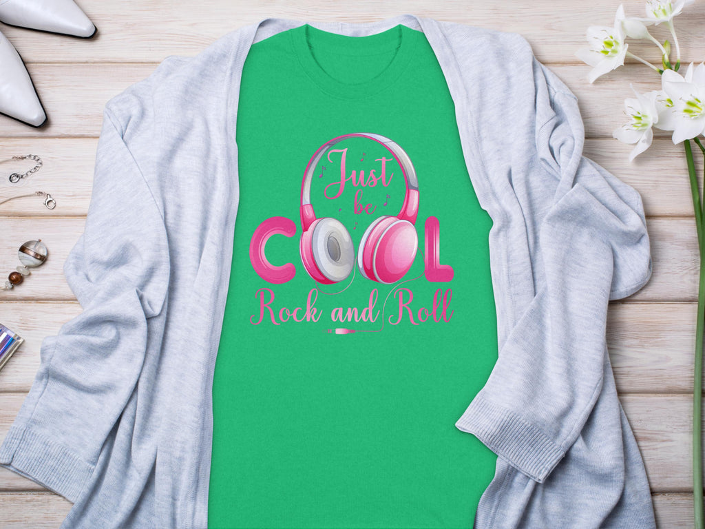 The Garment Graphics green T-shirt, perfect for music lovers, features Just be cool, Rock and Roll! with pink and white headphones. Its styled on wood with a light gray cardigan, accompanied by white shoes, jewelry, and white flowers.