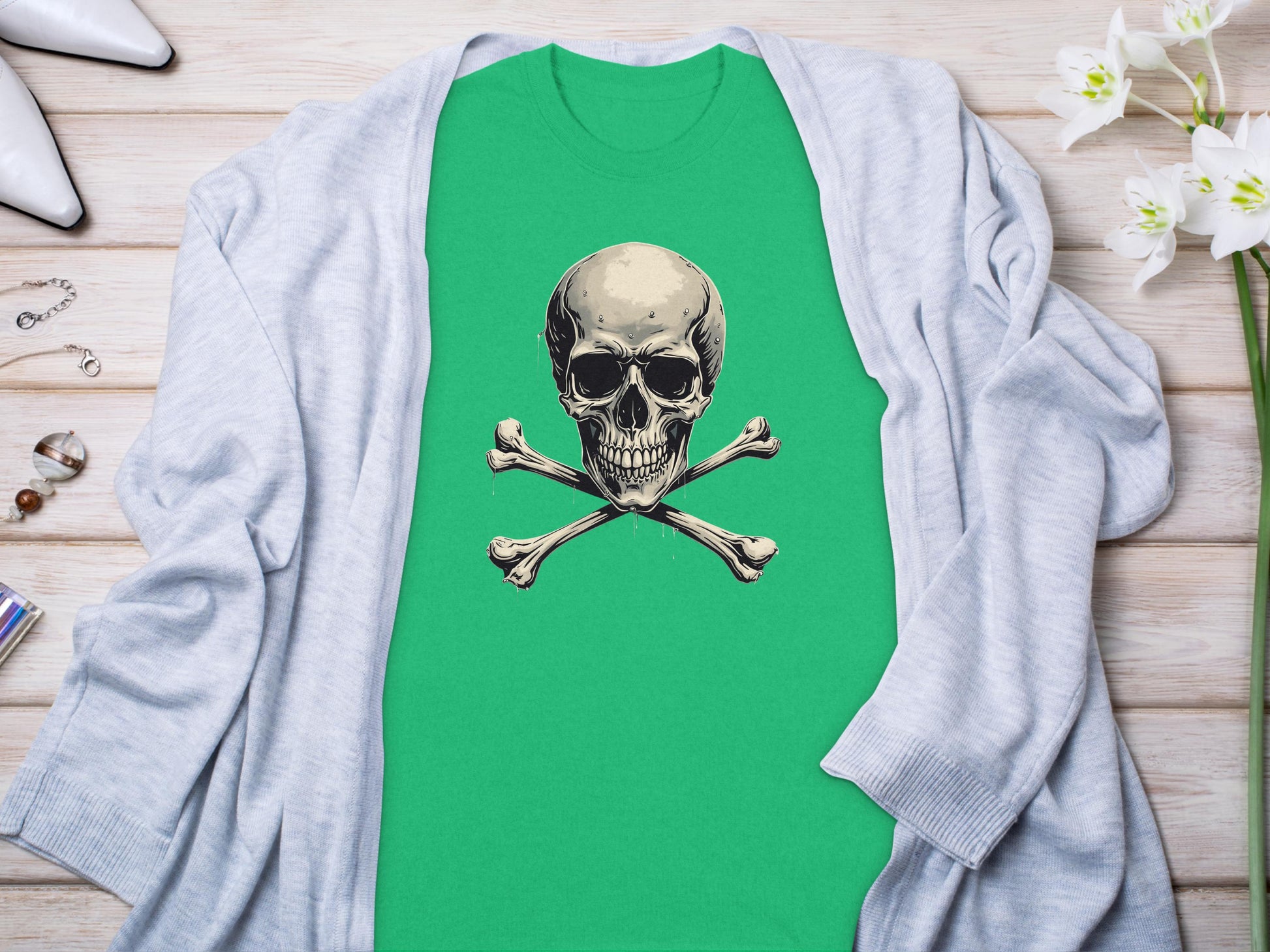 A green Garment Graphics graphic t-shirt with a striking skull and crossbones design is laid on wood. Its stylishly paired with a gray cardigan, white high heels, jewelry, and delicate white flowers for an edgy fashion look.