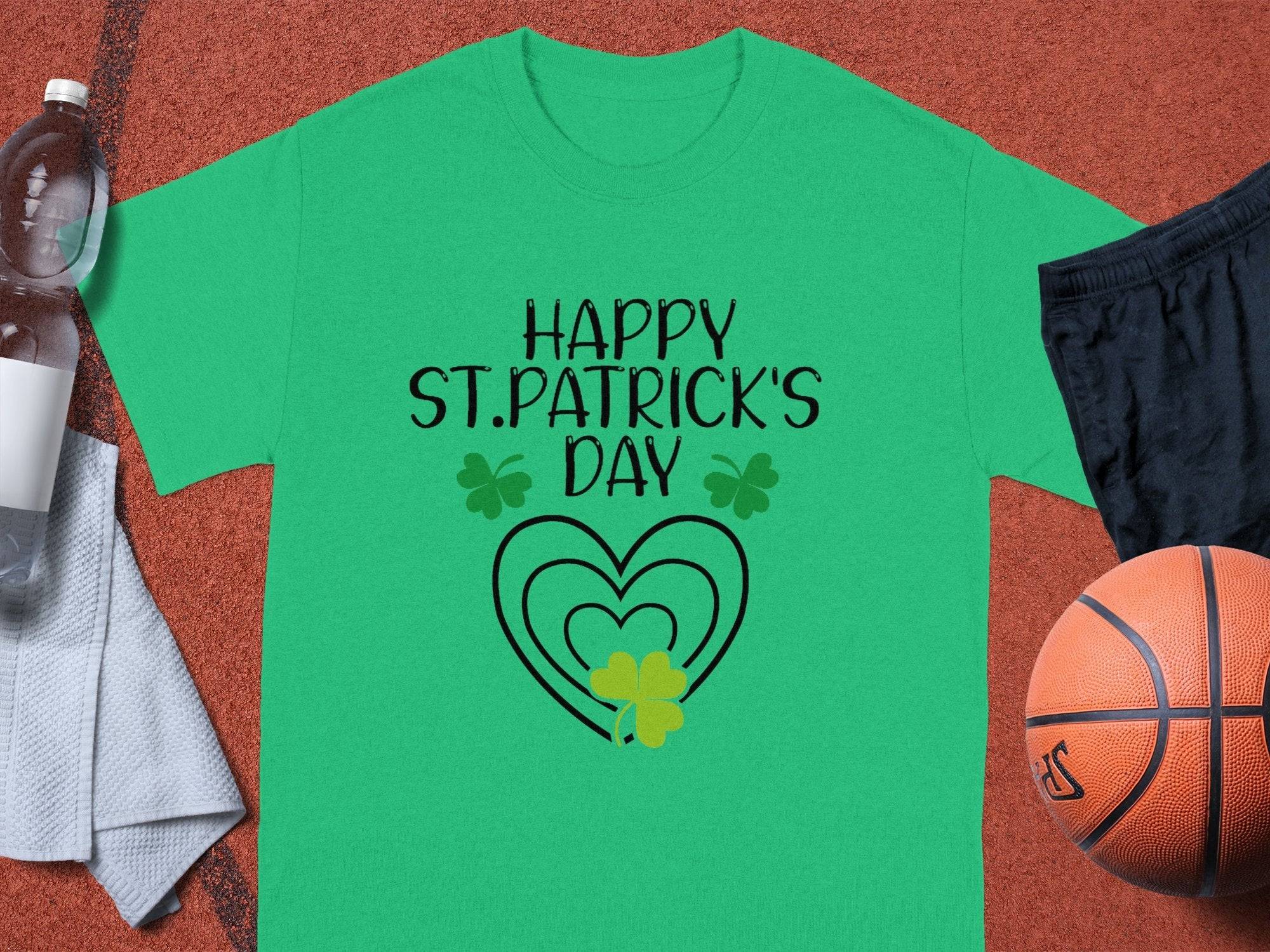 Garment Graphics T-shirt with HAPPY ST. PATRICKS DAY above a heart and shamrock adds festive flair, surrounded by a water bottle, towel, black shorts, and an orange basketball on a textured surface.