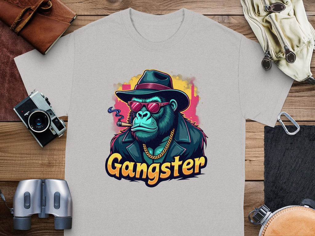 Garment Graphics Gangster Gorilla T-Shirt features a classic fit and urban style with a grey gorilla design adorned in a hat, sunglasses, and gold chain. The word Gangster adds flair, complemented by surrounding camera gear, binoculars, and leather items on wooden surface.