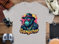 Garment Graphics Gangster Gorilla T-Shirt features a classic fit and urban style with a grey gorilla design adorned in a hat, sunglasses, and gold chain. The word Gangster adds flair, complemented by surrounding camera gear, binoculars, and leather items on wooden surface.