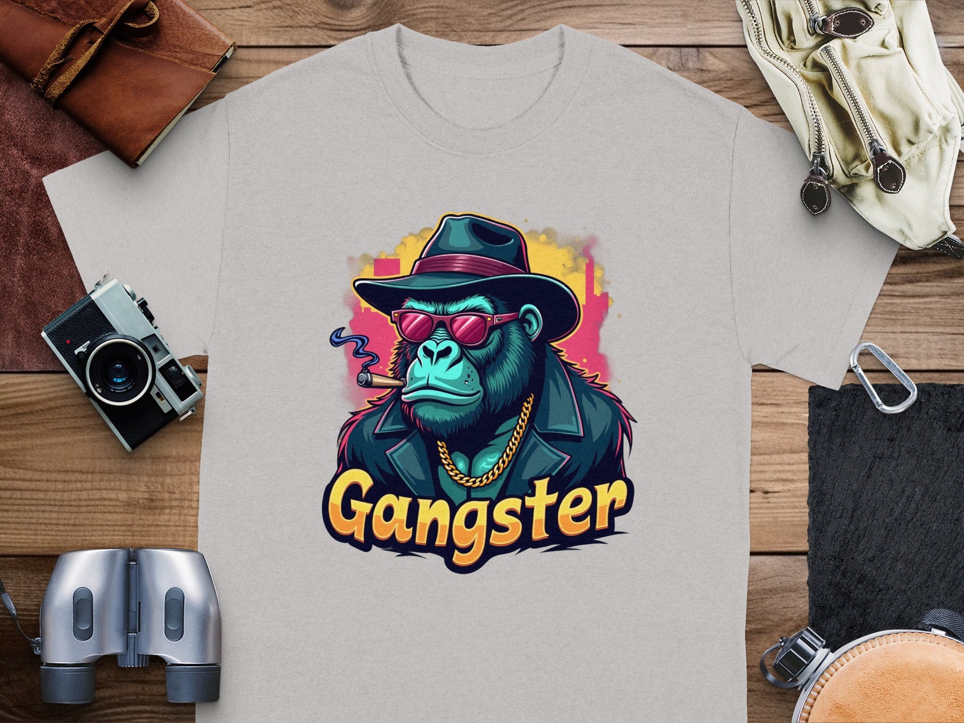 Garment Graphics Gangster Gorilla T-Shirt features a classic fit and urban style with a grey gorilla design adorned in a hat, sunglasses, and gold chain. The word Gangster adds flair, complemented by surrounding camera gear, binoculars, and leather items on wooden surface.