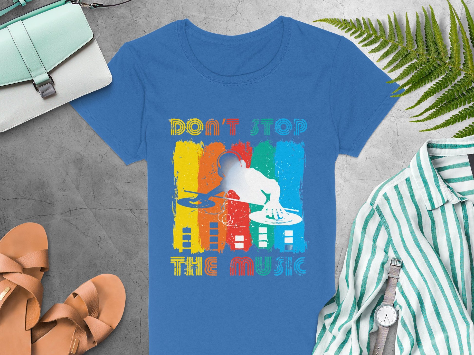The Garment Graphics blue DJ graphic tee showcases a vibrant DJ silhouette with DONT STOP THE MUSIC text—ideal for music enthusiasts. Its paired with items like a striped shirt, sandals, a wristwatch, a light blue handbag, and green ferns on a gray textured backdrop.