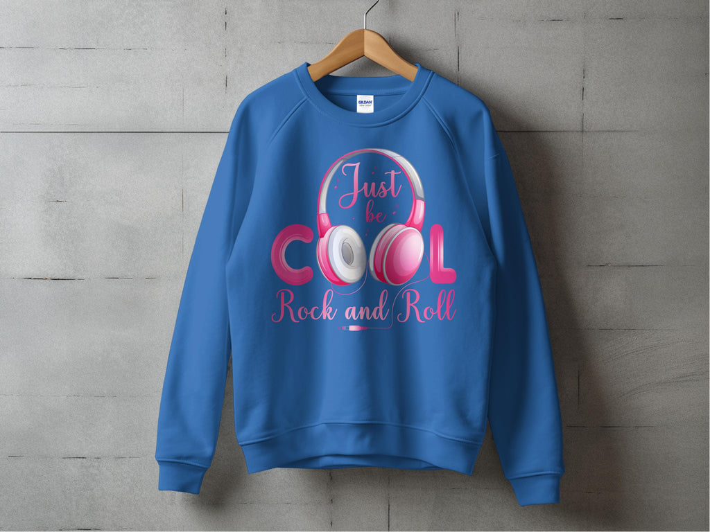 A blue sweatshirt by Garment Graphics hangs on a wooden hanger against a concrete wall, ideal for fashion and music lovers. It features white and pink rock and roll headphones with Just be Cool Rock and Roll in pink and white letters.