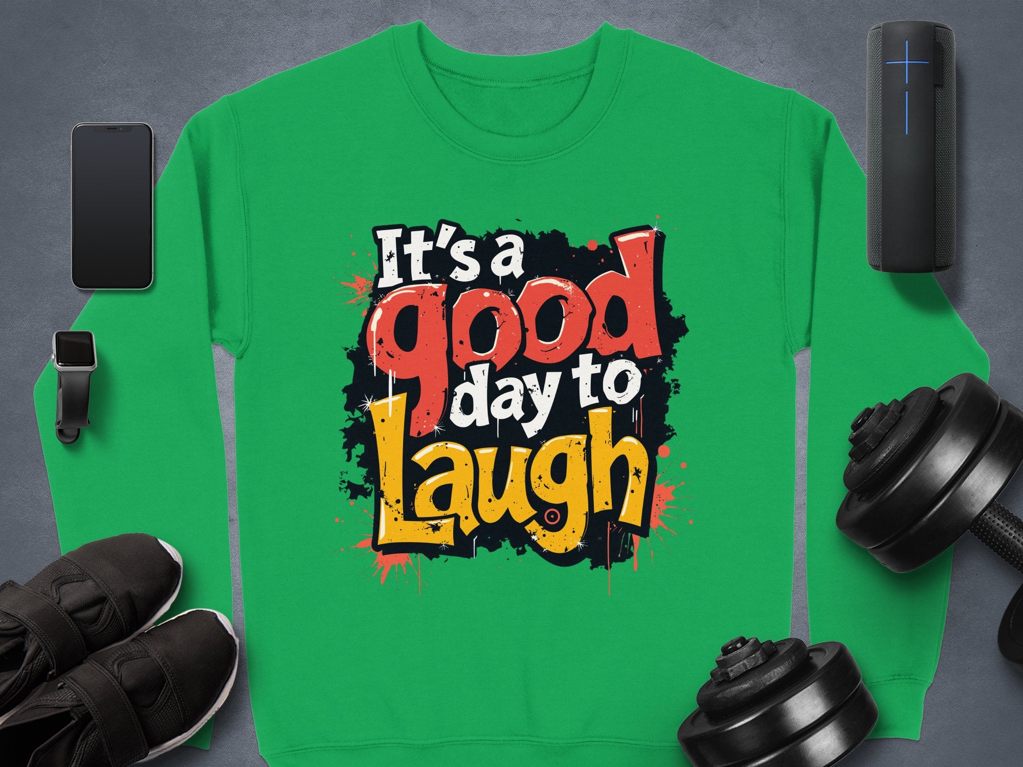 A Garment Graphics medium-weight unisex sweatshirt with Its a good day to Laugh in bold, colorful print, surrounded by a smartphone, smartwatch, speaker, black shoes, and dumbbells on a dark gray background.