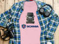 The Garment Graphics pink Scania T-shirt with a truck graphic, great for automotive fans, lies over a blue plaid shirt. A DSLR camera, headphones, and a coffee cup lid rest nearby on the wooden surface, crafting an inviting setting for graphic T-shirt lovers.