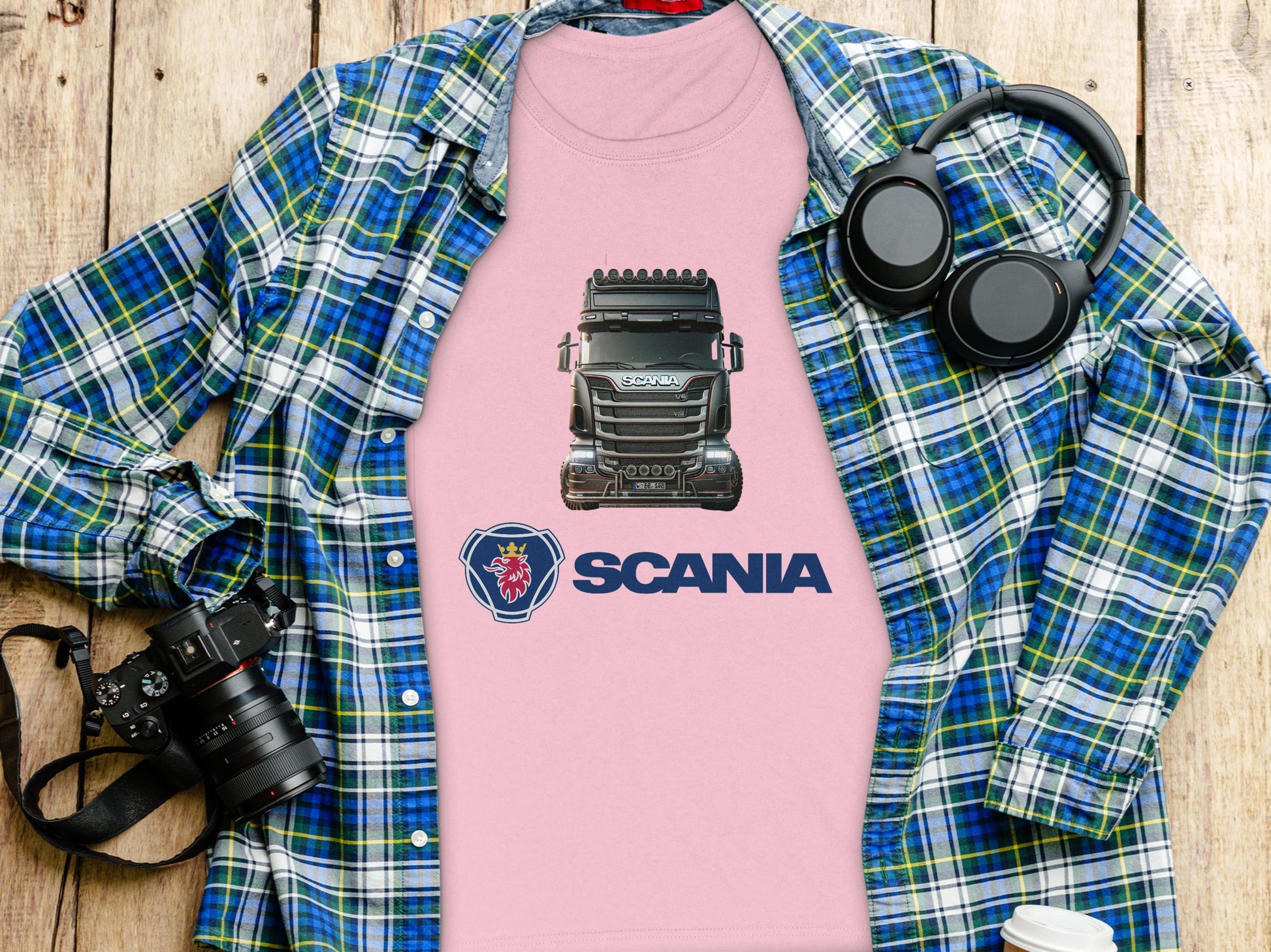 The Garment Graphics pink Scania T-shirt with a truck graphic, great for automotive fans, lies over a blue plaid shirt. A DSLR camera, headphones, and a coffee cup lid rest nearby on the wooden surface, crafting an inviting setting for graphic T-shirt lovers.