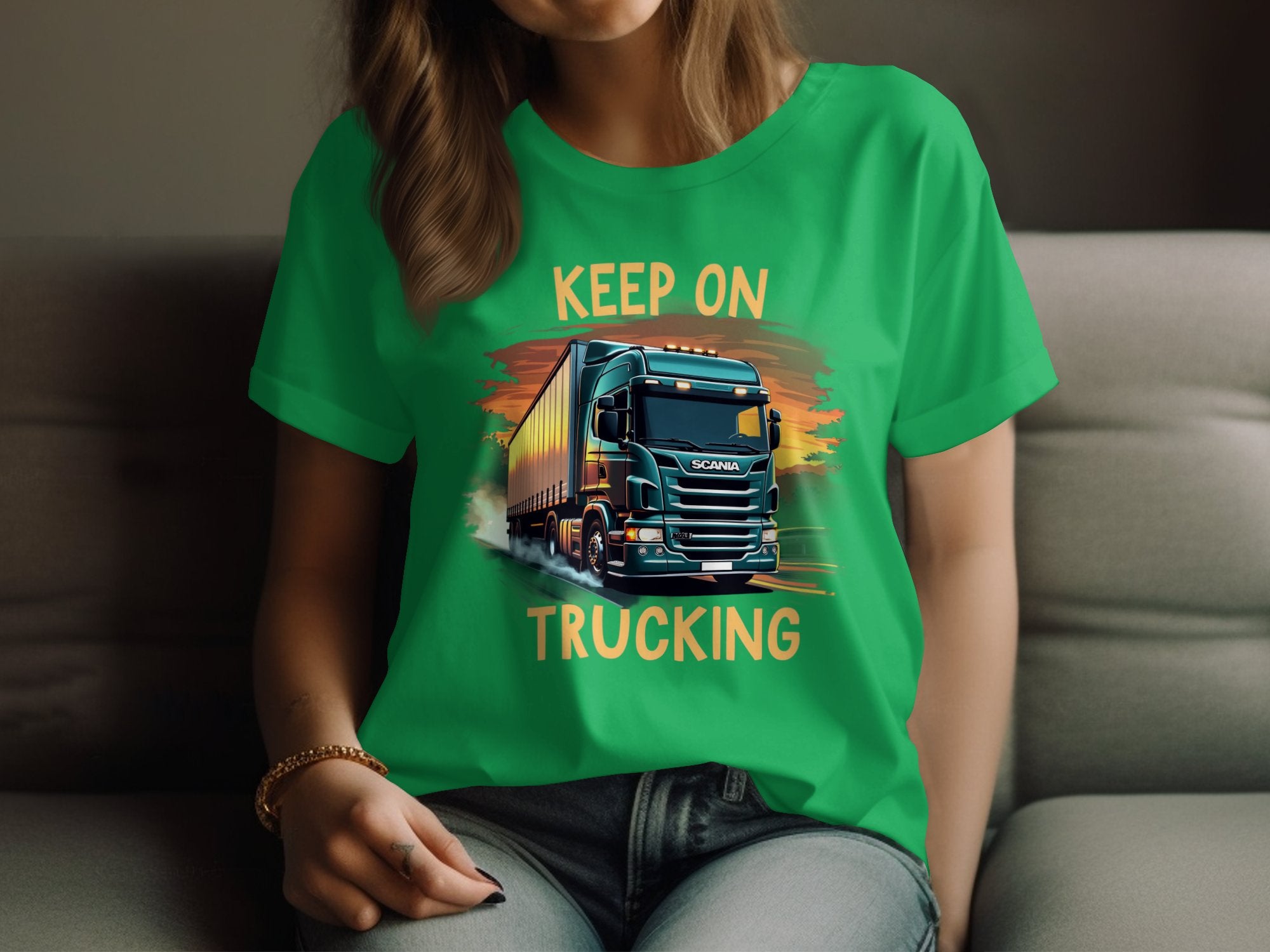 A person wears a Garment Graphics green T-shirt, featuring \KEEP ON TRUCKING\ and a truck graphic. This stylish piece, favored by trucking enthusiasts, showcases dynamic colors that bring the illustration to life as they sit on a couch.