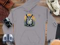 Introducing Garment Graphics Fierce Owl Illustration Hoodie, made from medium-heavy fabric. This gray hoodie boasts a unique owl graphic with large eyes on a yellow-orange background, styled on wood with adventure gear like a camera and binoculars.