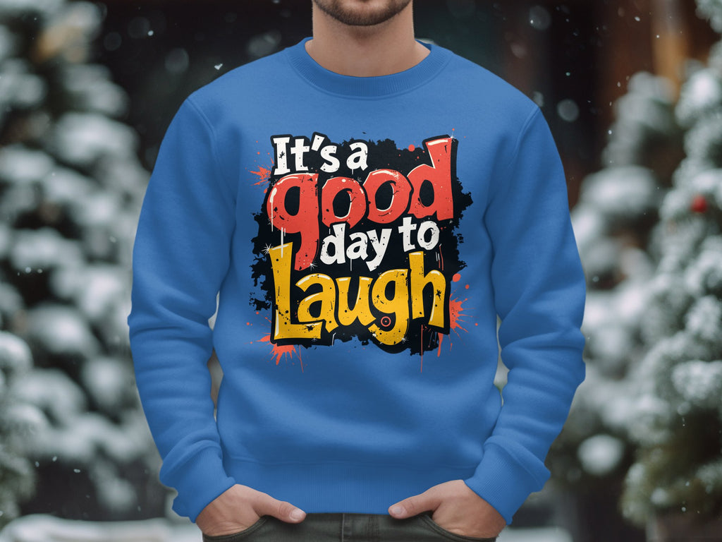 Outdoors, a person wearing a Garment Graphics blue graphic art sweatshirt stands amidst snow-covered trees and blurred lights. The bold, colorful text on the unisex-sized garment reads Its a Good Day to Laugh, perfectly capturing the serene moment.