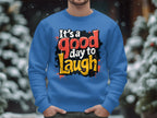 Outdoors, a person wearing a Garment Graphics blue graphic art sweatshirt stands amidst snow-covered trees and blurred lights. The bold, colorful text on the unisex-sized garment reads Its a Good Day to Laugh, perfectly capturing the serene moment.