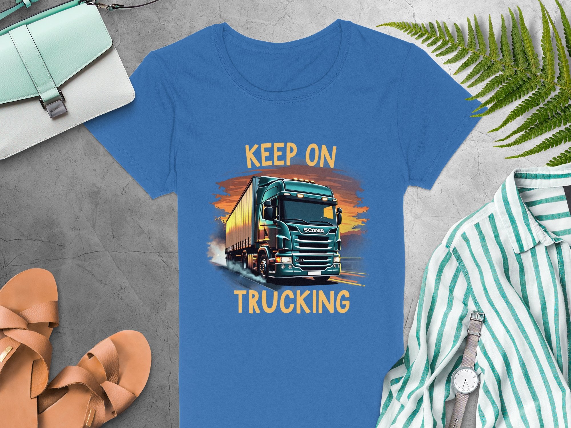 The Keep on Trucking T-shirt by Garment Graphics, ideal for truck lovers, showcases a lively truck graphic at sunset. Styled on a gray background with a green striped shirt, sandals, purse, watch, and fern leaves for an adventurous and relaxed vibe.