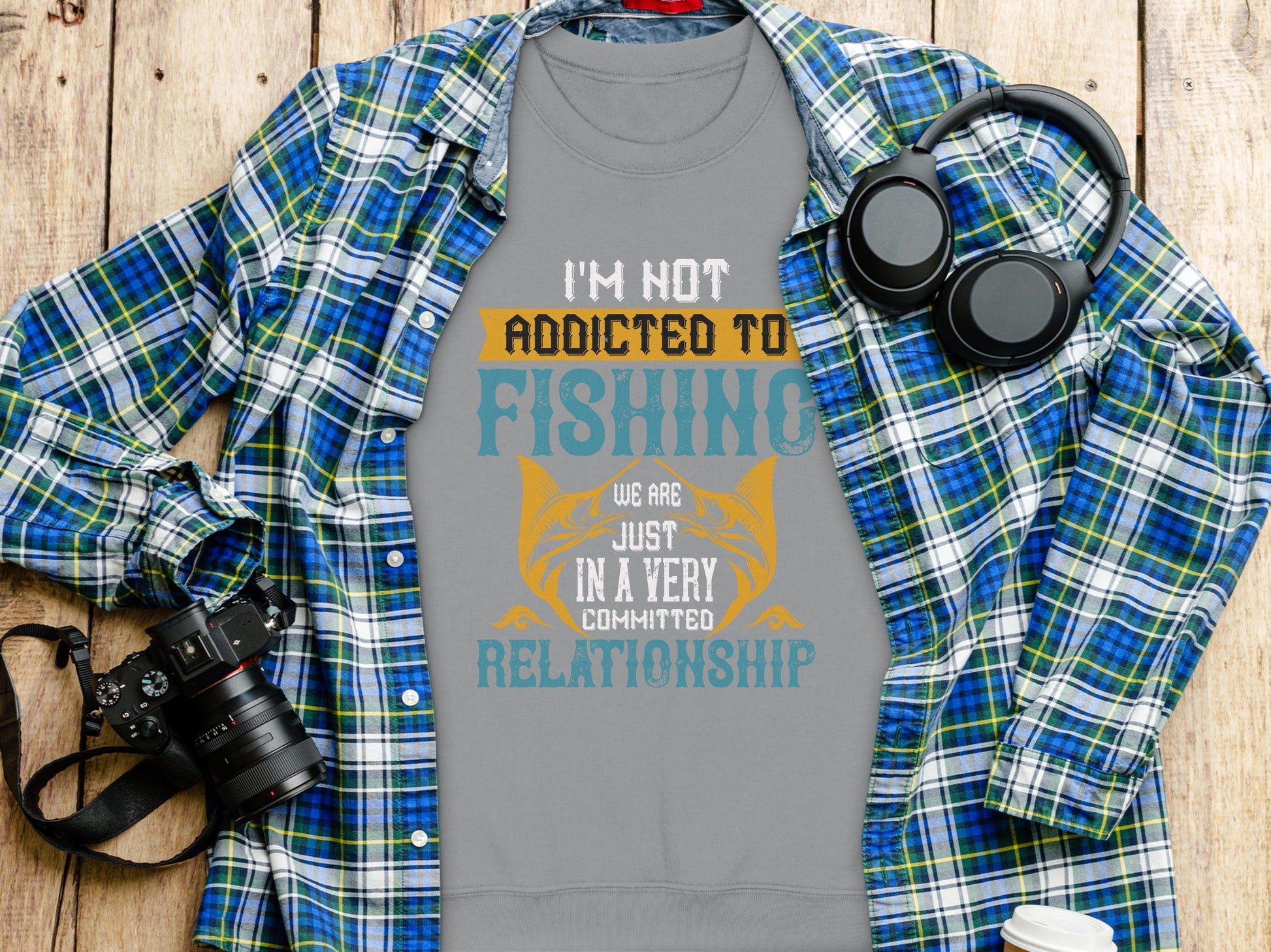 A Garment Graphics gray unisex t-shirt with the text Im not addicted to fishing, we are just in a very committed relationship lies on a wooden surface alongside a blue plaid shirt, headphones, a camera, and a disposable cup.