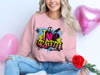 A person sits cross-legged with a red rose, wearing a Garment Graphics sweatshirt featuring a graffiti design that reads I ❤️ GRAFFITI. Blue ripped jeans complete the urban style, with two pink heart-shaped balloons floating in the background.