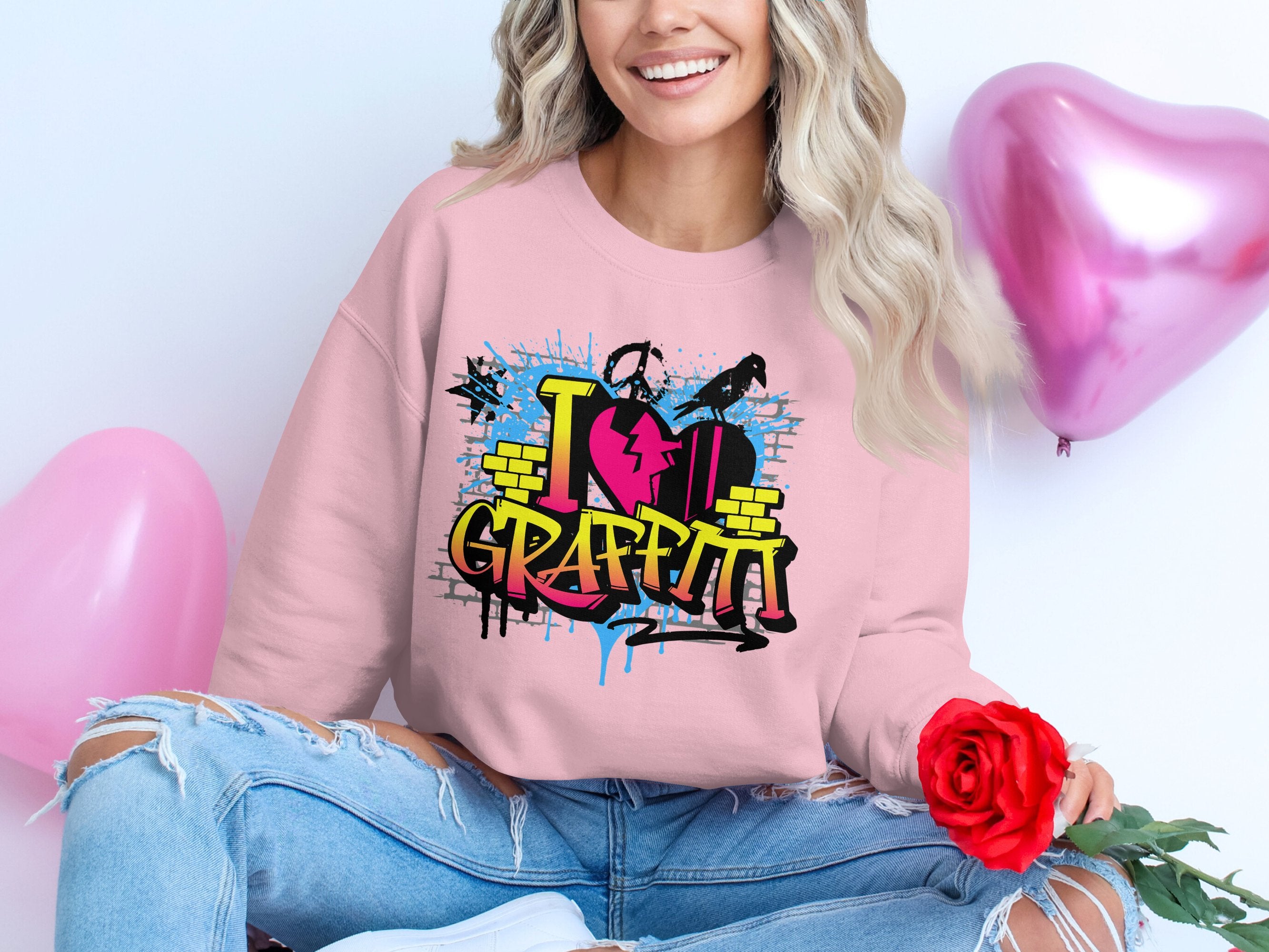 A person sits cross-legged with a red rose, wearing a Garment Graphics sweatshirt featuring a graffiti design that reads I ❤️ GRAFFITI. Blue ripped jeans complete the urban style, with two pink heart-shaped balloons floating in the background.